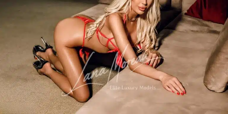 High-class escort in red lingerie posing seductively on a luxury sofa